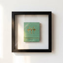 Framed print of The Happiest Hour Matchbox hanging on a white wall with cozy warm lighting, showcasing glossy and matte textured paper in an inviting setting.