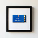 Framed Little Prince print on white wall, black wood frame, natural daylight, cozy environment.