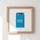 Full shot of 'Uptown Social' framed print on white wall, featuring textured paper and natural wood frame with warm natural window light, in a cozy setting.